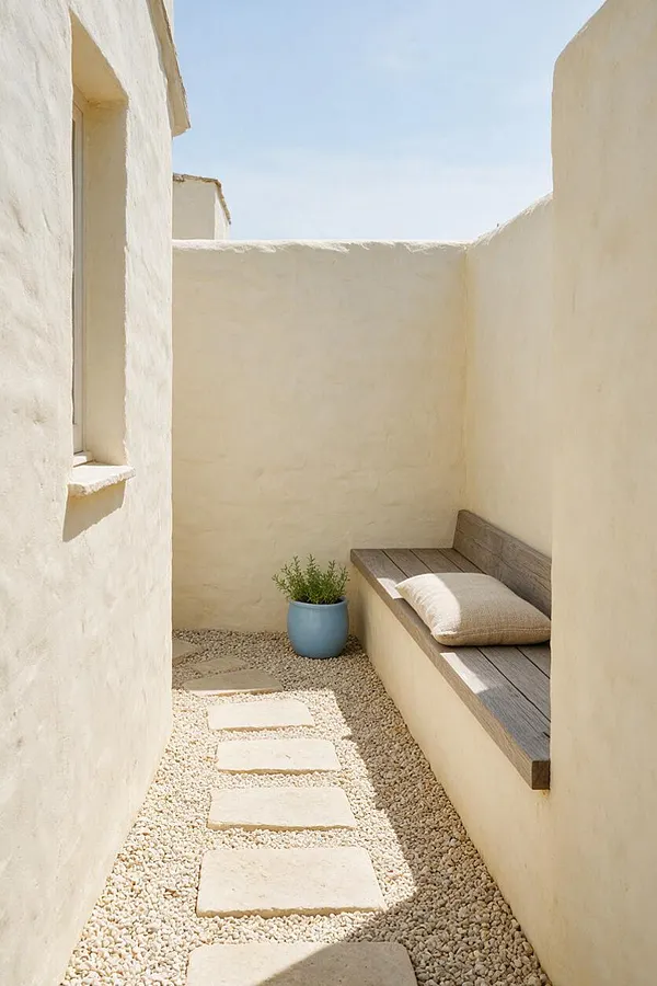 A narrow coastal courtyard with creamy stucco walls, a pebble ground cover, and a stepping-stone path leading to a built-in wooden bench. A soft cushion and a blue planter with greenery add a relaxed, airy feel under bright natural light.