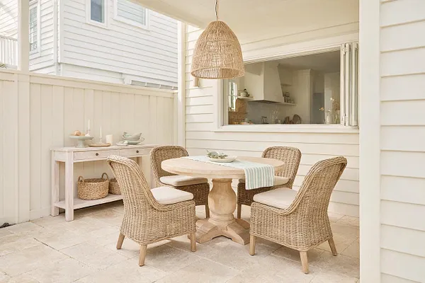 A bright backyard dining nook with a round pedestal table and four woven wicker chairs on a pale stone patio. White clapboard siding, a matching fence, and a large woven pendant light create a relaxed coastal feel, with a simple side console and soft neutral decor completing the space.