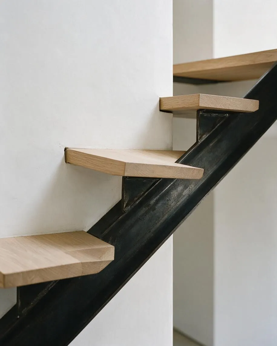 A close-up of minimalist floating stairs with light oak wood treads mounted to a black steel stringer. The stairs rise beside smooth white walls, creating a clean contrast of natural wood, dark metal, and bright neutral surfaces.