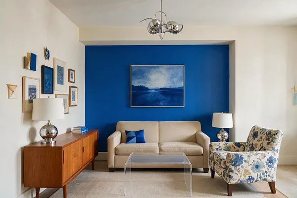 A bright eclectic living room with a deep cobalt blue accent wall framing a cream sofa, a floral upholstered armchair, and a clear acrylic coffee table. Framed artwork, a walnut sideboard, chrome lamps, and a modern ceiling fixture add a mix of vintage and contemporary details.