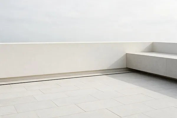 A sparse minimalist rooftop terrace with large pale tiled flooring, a low white perimeter wall, and a built-in bench or ledge in light gray on the right. The space is open to a bright sky, with soft shadows emphasizing the simple geometry and uncluttered design.