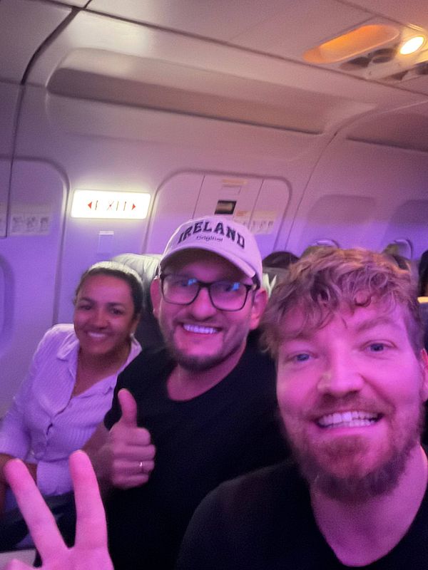 A man wearing a blue shirt and cap is sitting in an airplane seat, drinking from a green cup.