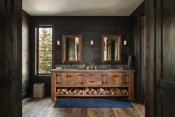 A rustic powder room with dark wood plank walls, a long reclaimed wood double vanity, and two framed mirrors above a stone-toned countertop. Firewood is stacked on an open shelf beneath the sink, with a window on the left bringing in natural light and a blue rug softening the wood floor.