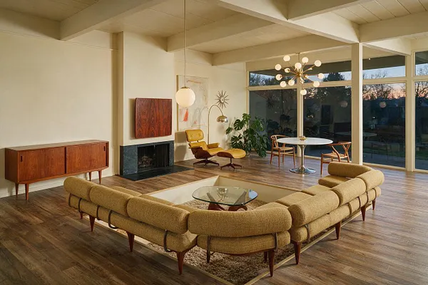 A spacious mid-century modern game room with a sectional curved around a square glass coffee table, walnut cabinetry, a black fireplace, and a yellow lounge chair. Large floor-to-ceiling windows, a round pedestal table, and an atomic-style chandelier create an open, retro-inspired atmosphere.