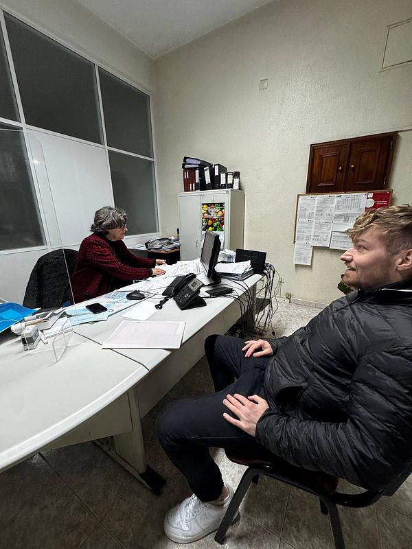 A man is seated in an office while a woman works at a desk, likely discussing driving school enrollment.