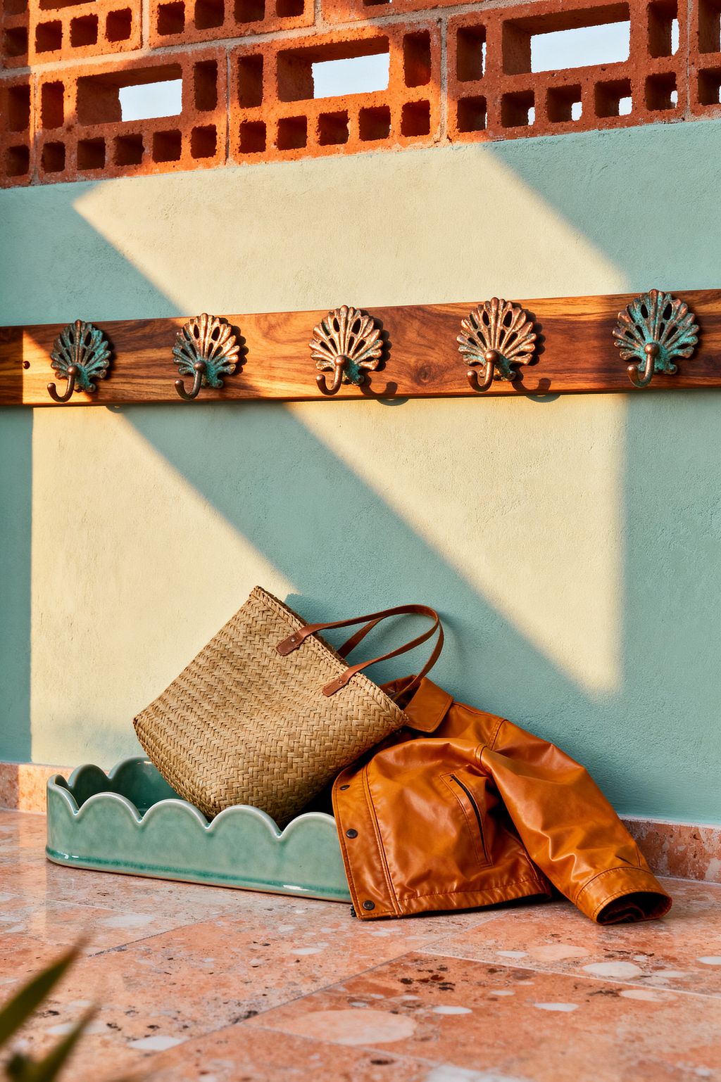 A tropical mudroom vignette features a seafoam plaster wall with a teak rail of verdigris, palm-shaped hooks beneath a terracotta breeze-block screen that filters sun into diagonal shadows. On the peach terrazzo floor, a scalloped green glazed ceramic tray holds a woven rattan tote while a caramel leather jacket rests beside it, blending cool greens with warm brick, wood, and leather for a breezy, coastal mood.
