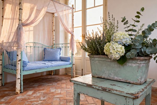 A sunlit patio in shabby chic style features a distressed mint-green metal daybed framed by birch trunks and draped with sheer linen, topped with blue-and-white gingham cushions and bolsters. In the foreground, a weathered mint table supports a patinated planter filled with pale hydrangeas, lavender, eucalyptus, and herbs, while terracotta herringbone brick flooring and French doors with brass hardware glow in warm evening light.