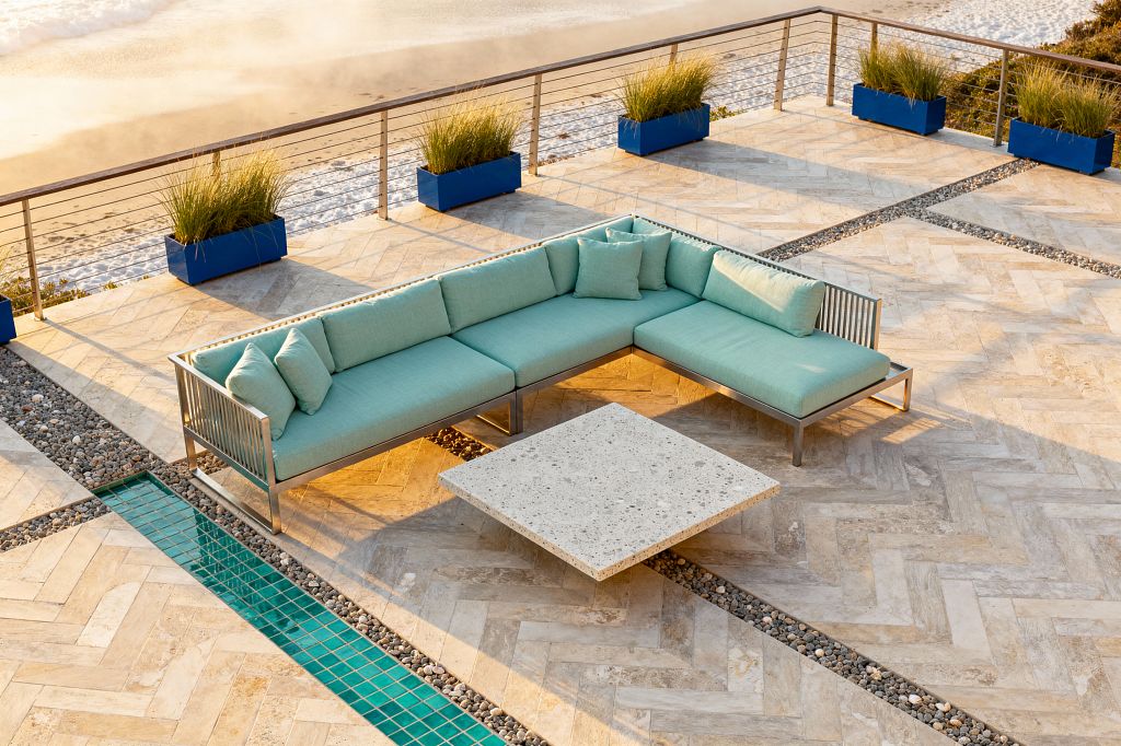 A beachside patio features an L-shaped outdoor sectional with seafoam cushions on a brushed aluminum frame around a low square terrazzo coffee table. The flooring is sandy beige travertine in a herringbone pattern with river-pebble insets and a narrow run of teal glass tiles. Cobalt blue planters with grasses line a stainless cable railing overlooking sand and surf, while warm sunlight casts long shadows across the scene.