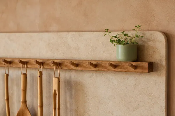 A biophilic-style wall detail features a warm oak peg rail mounted on a pale stone or plaster surface. Bamboo kitchen tools hang below, while a small sage-green planter with trailing greenery sits on the right end of the rail.