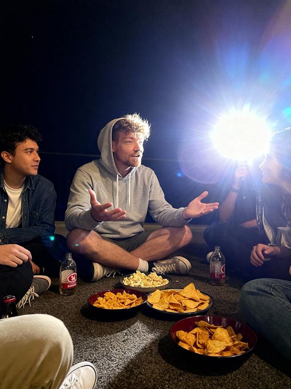 A group of friends is sitting on the floor, enjoying snacks and engaging in conversation under a bright light.