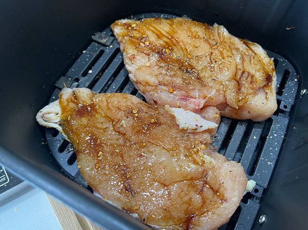 Two marinated chicken breasts are placed in an air fryer basket.