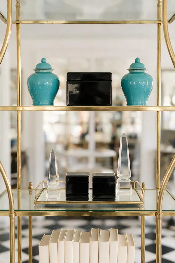 A Hollywood Regency-style etagere with a polished brass frame and glass shelves displays glossy turquoise jars, black lacquer boxes, clear crystal obelisks, and cream books. The blurred background and black-and-white checkered floor add depth and classic glamour.