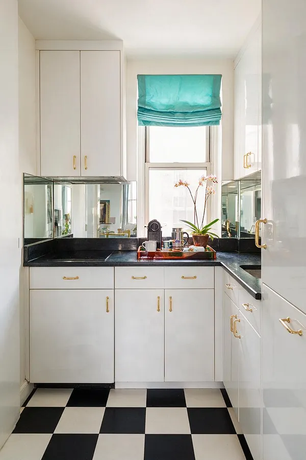 A narrow studio apartment kitchen with glossy white cabinets, gold hardware, black countertops, and mirrored backsplash panels on both sides. A small window with a turquoise Roman shade brightens the space, while a black-and-white checkerboard floor adds bold Hollywood Regency flair.