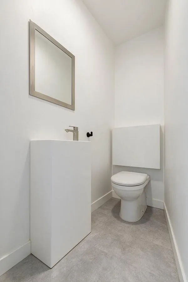 A compact powder room with white walls, a slim framed mirror, a minimalist white vanity with a simple chrome faucet, and a wall-mounted toilet. The space has light gray flooring and a clean, uncluttered modern look.