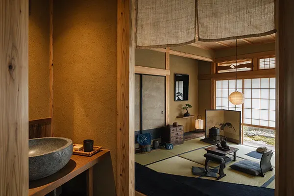 A Japanese Zen-style powder room with a stone vessel sink on a wooden ledge, textured plaster walls, and warm timber framing. Beyond the entry, a traditional tatami room with shoji screens, low seating, paper lantern lighting, and a calm neutral palette creates a peaceful atmosphere.