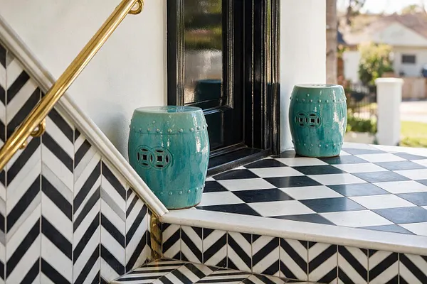 An exterior porch in Hollywood Regency style featuring a black-and-white checkerboard floor, bold chevron tile risers, glossy black front door, and a brass handrail. Two turquoise ceramic garden stools add color and a glamorous decorative accent.
