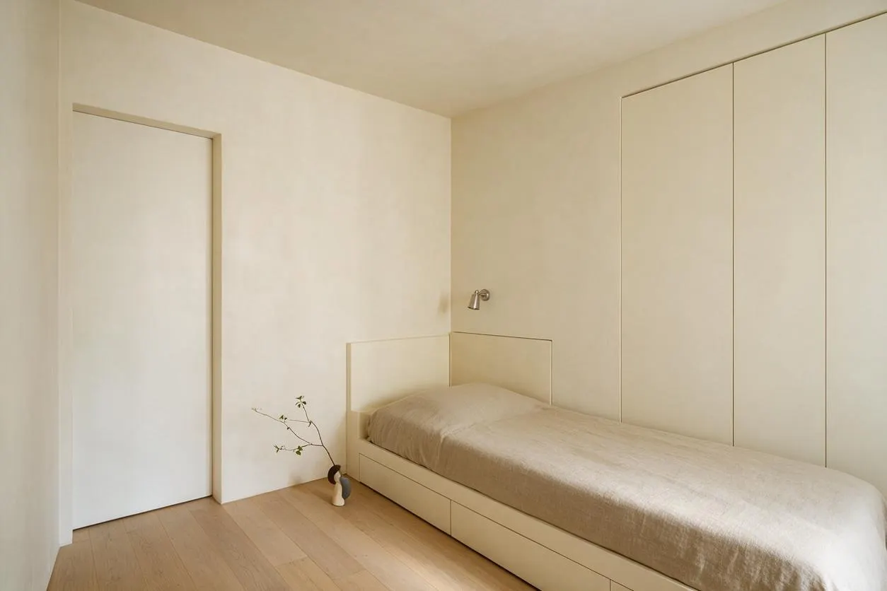 A minimalist guest bedroom with a single low bed dressed in beige bedding, built-in paneled storage along the wall, and a small wall-mounted reading light above the headboard. The room has white walls, light wood flooring, a plain door, and a simple vase with a bare branch adding a subtle decorative touch.