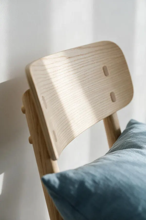 A close-up of a Scandinavian-style wooden chair with a softly curved light ash backrest and simple joinery details. A muted blue cushion or pillow rests against the seat, while sunlight casts gentle shadows on a white wall behind it.