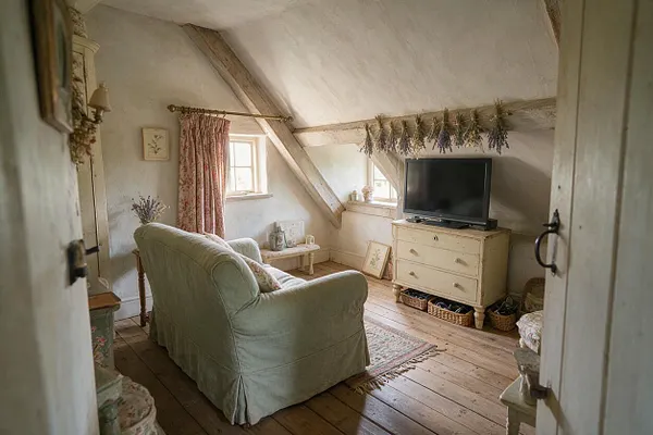 A cozy attic media room with sloped whitewashed walls, exposed beams, and a soft sage slipcovered loveseat facing a flat-screen TV on a cream painted dresser. Pink floral curtains, dried lavender garlands, woven baskets, and rustic wood floors create a warm cottagecore atmosphere.