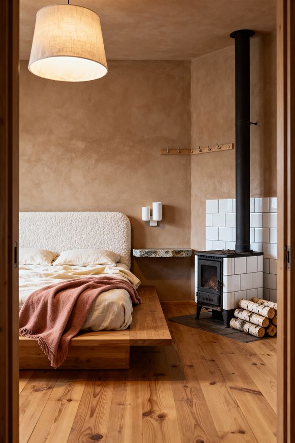 Cozy Scandinavian bedroom seen through a doorway. A low pine platform bed with an off-white bouclé headboard sits against clay-beige plaster walls. Ivory linen bedding is draped with a dusty rose throw. To the right, a compact cast-iron wood stove rises to a black flue, set within glossy white square tiles; stacked birch logs rest beside it. A floating stone night shelf, a simple peg rail, warm wood plank flooring, and a linen drum pendant complete the serene space.