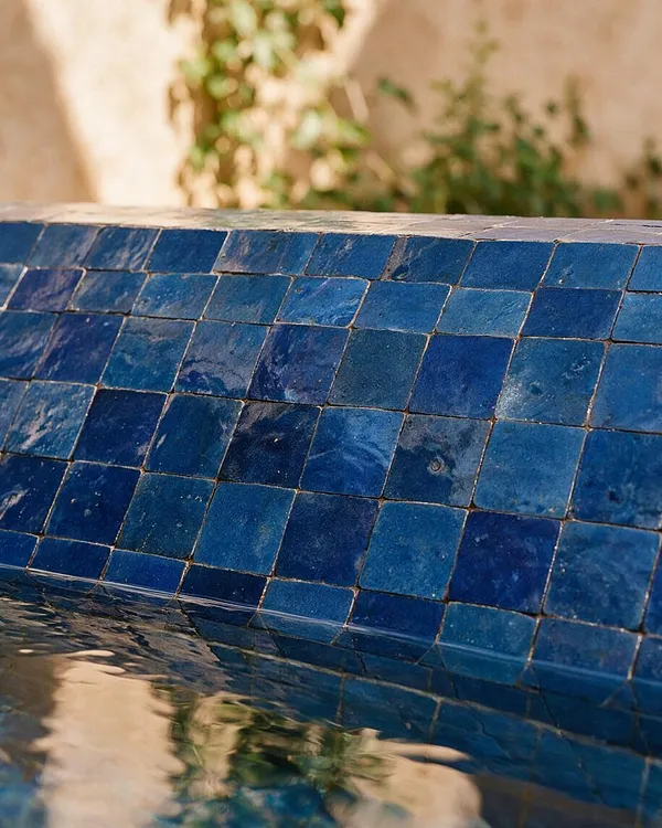 A close-up of a curved pool edge covered in small square blue Moroccan-style tiles with varied glossy finishes. The water below reflects the tiles and surrounding greenery against a warm, blurred background.