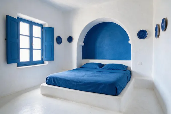 A bright Mediterranean-style guest bedroom with white stucco walls, a built-in bed platform, and a cobalt blue arched alcove behind the headboard. Matching blue bedding and a blue shuttered window create a crisp, sunlit contrast, while small blue wall plates add simple decorative detail.