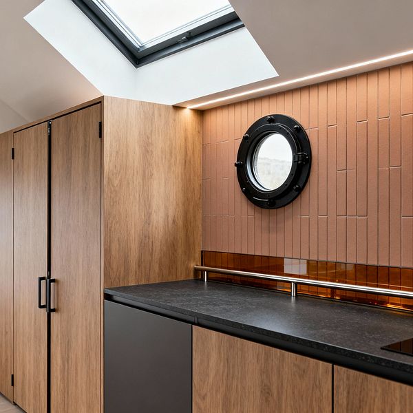 A modern laundry room features flat-front oak cabinets and matte charcoal base units topped with a dark stone countertop. A wall of narrow vertical terracotta tiles is accented by an amber glass strip and slim stainless rail, with a round black-framed porthole window at center; recessed linear LED lighting and a large skylight in the sloped ceiling brighten the space and highlight its clean lines and warm, earthy palette.