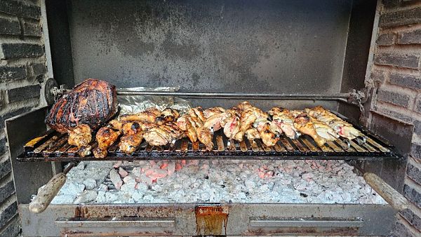 Close-up view of a BBQ grill loaded with various meats including sausages, pork chops, and chicken wings, surrounded by a plate of salad and a wooden table setting.