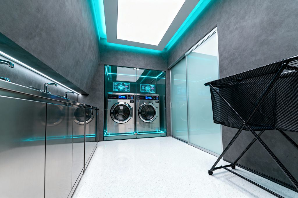 A sleek laundry room shows two stainless front-loading washers enclosed behind glass, outlined by cyan LED lighting with digital displays above. On the left, reflective stainless cabinets with integrated sinks and under-cabinet lighting run the length of the wall; on the right, a frosted glass sliding door and a black mesh rolling hamper sit on a bright white speckled floor beneath a large recessed ceiling light and turquoise cove glow.