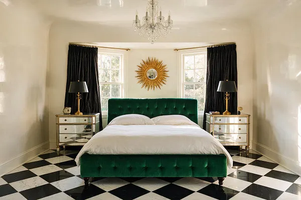 A Hollywood Regency master bedroom with a deep green velvet bed centered between two mirrored nightstands. Black drapes frame bright windows behind a gold sunburst mirror, while a crystal chandelier hangs overhead and a black-and-white checkerboard floor adds bold contrast.
