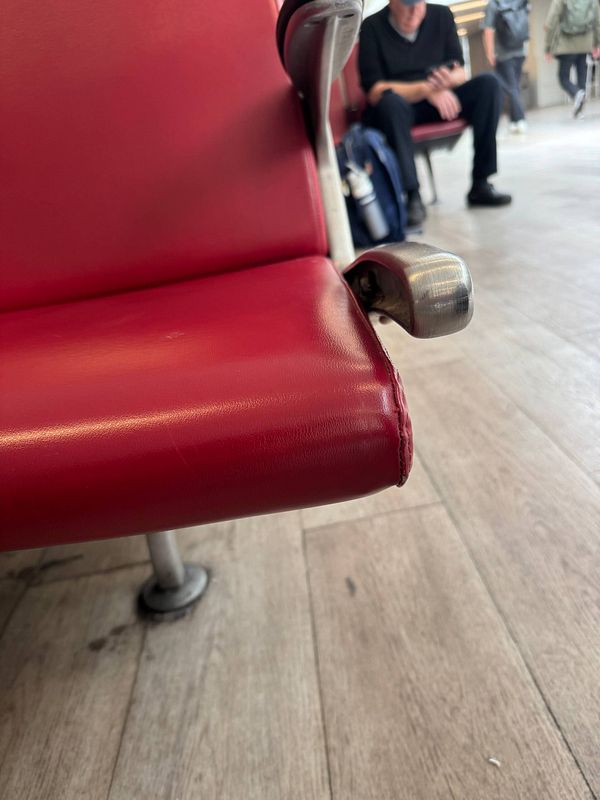 A close-up view of a worn red airport seat with visible damage and a busy terminal background.