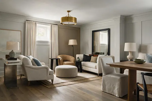 A transitional basement living area with soft beige and cream seating arranged around a textured rug, a round ottoman, and black accent tables. The room features wood floors, a gold drum ceiling light, tall curtains, paneled walls, a large mirror, and a dining nook with a wood table and upholstered chairs.