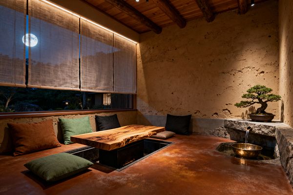 An intimate Japanese-style dining room features a live-edge wood table over a recessed leg well, with low floor cushions in green, rust, charcoal, and taupe along clay plaster walls. Warm cove lighting above sheer linen window shades and a spotlight on a bonsai create calm ambiance, while a rough stone ledge with a brass spout pours into a metal bowl fountain; the burnished red earthen floor and exposed timber ceiling beams emphasize natural textures, with a moonlit garden visible outside.