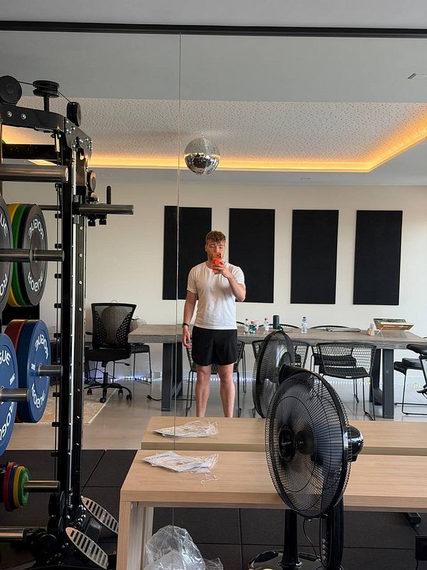 Photo of a young man with a light blue towel around his neck posing for a gym selfie with exercise machines in the background.