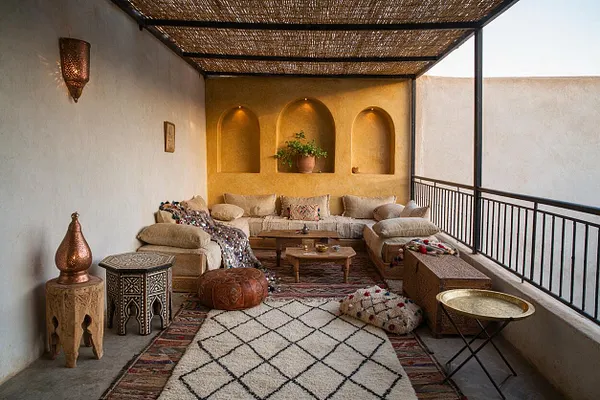 A covered front porch styled as a Moroccan-inspired lounge with low beige sectional seating, layered patterned rugs, carved side tables, and brass-toned decor. Built-in arched wall niches and a woven reed ceiling add texture, while white plaster walls and black metal railings frame the cozy outdoor space.