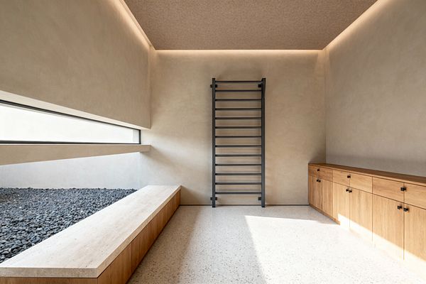 A minimalist home gym centers a black wall-mounted stall bar on a warm beige plaster wall, with a long run of natural oak cabinets with black pulls to the right and a low bench topped with light travertine beside a recessed bed of gray river pebbles to the left. A slim horizontal window and perimeter cove lighting brighten the pale terrazzo floor, emphasizing clean lines and an uncluttered layout suited to stretching and body-weight training.