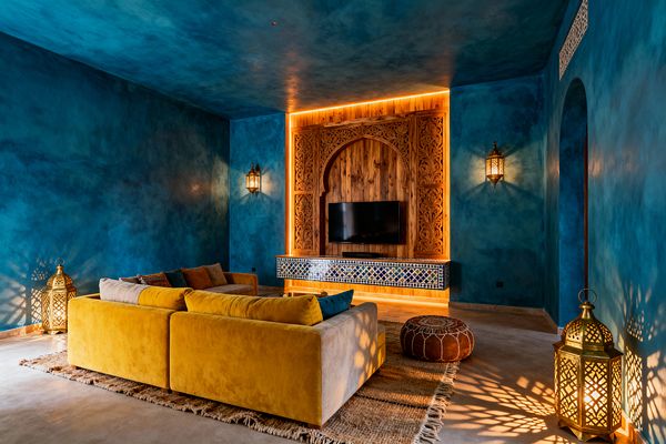 A Moroccan-style media room features deep cobalt-blue tadelakt walls and ceiling surrounding a carved wood arched TV wall with warm LED backlighting and a blue-and-white zellige tile console. A mustard velvet sectional sits on a fringed jute rug beside a cognac leather pouf, while brass pierced lanterns and wall sconces cast patterned light. The room includes an arched doorway and smooth neutral flooring, creating a cinematic, intimate atmosphere.