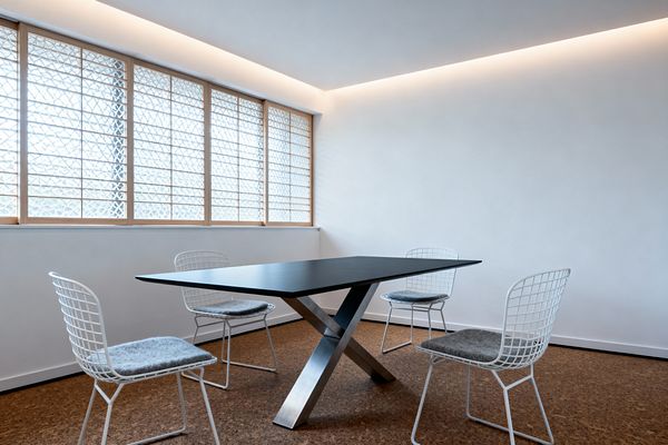 A minimalist dining room features a rectangular black table with an X-shaped brushed steel base, surrounded by four white wire chairs with gray felt cushions. Warm cork flooring contrasts with crisp white walls and a continuous cove of concealed LED lighting that washes the ceiling. A wall of windows with light natural wood frames and patterned translucent glass softly filters daylight, keeping the space bright yet private. The room is uncluttered, emphasizing clean lines and a calm, airy atmosphere.