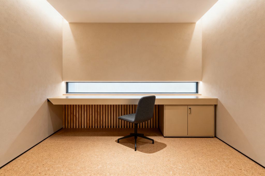A pared-back home office with warm beige plaster walls and a long built-in desk spanning the width of the room. A narrow horizontal frosted window runs above the work surface, washing the space with diffuse light. Vertical oak slats form a panel beneath the desk, while a sand-colored cabinet sits to the right. The floor is cork, and a charcoal upholstered swivel chair with a black metal base stands centered.