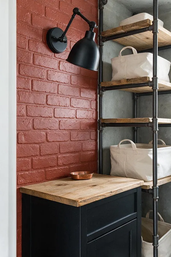 A compact industrial powder room features a red painted brick accent wall, a black adjustable wall sconce, and a dark vanity topped with a thick natural wood counter. To the right, open shelving made from black metal pipe and wood holds canvas storage bins against a gray concrete wall.