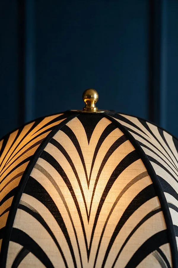 A close-up of an Art Deco-style lampshade with a cream fabric surface and bold black fan-like geometric stripes. A small polished brass finial sits at the top, and a deep blue background adds contrast behind the lamp detail.