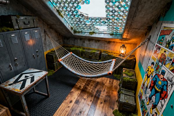 A kids’ room styled like a survival bunker featuring a rope hammock suspended between concrete walls above wood plank flooring and a black rubber mat. Gunmetal lockers and olive ammo boxes with patches of moss line the perimeter, a lantern glows on a crate, a sloped skylight patched with mosaic CDs filters blue light, and a retro comic-strip mural brightens a wall with teal and yellow stripes.