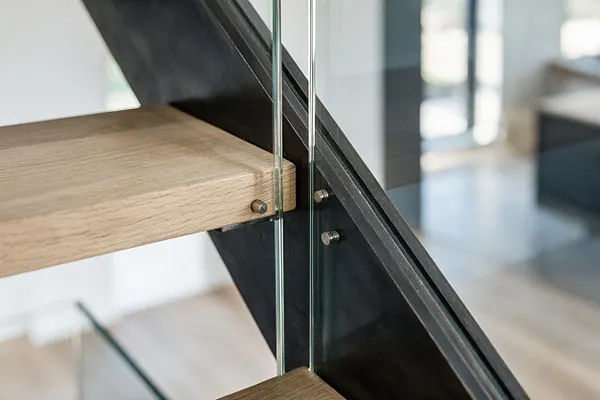 A close-up of a modern staircase showing a light oak wood tread attached to a black steel stringer with clear glass panels. Small metal bolts and a sleek diagonal support emphasize the clean, architectural construction.