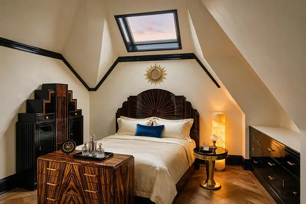 A compact guest bedroom with a dramatic Art Deco look, featuring a dark carved headboard, cream bedding, and black trim outlining angled walls and a skylight. Zebrawood-style furniture, gold accents, a glowing floor lamp, and a stepped cabinet create a luxurious, geometric atmosphere.
