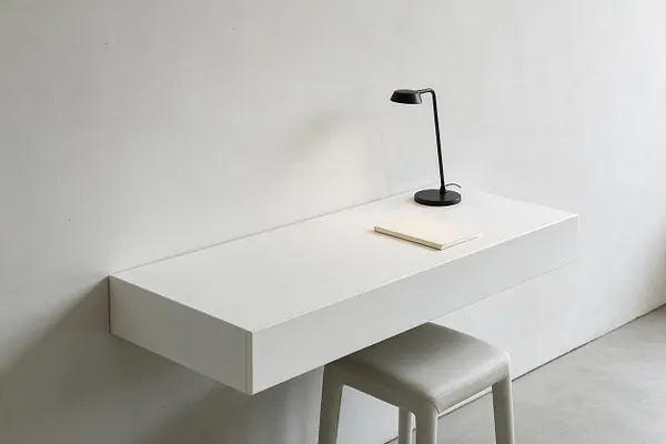 A minimalist workspace vignette shows a floating white wall-mounted desk against a plain white wall, with a small black task lamp and a slim notebook on top. A light gray stool tucks underneath, and the room is finished with a soft neutral palette and clean, modern lines.