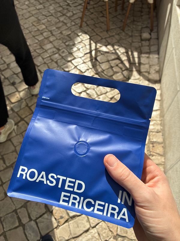 A hand holds a blue resealable coffee bag with white text on a cobblestone street background.