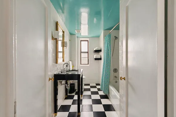 A narrow master bathroom with a glossy turquoise ceiling, black-and-white checkerboard floor, and white subway-tile walls. The room includes a black vanity with a marble top, brass-framed mirror and hardware, a small window, and a bathtub with a turquoise patterned shower curtain.