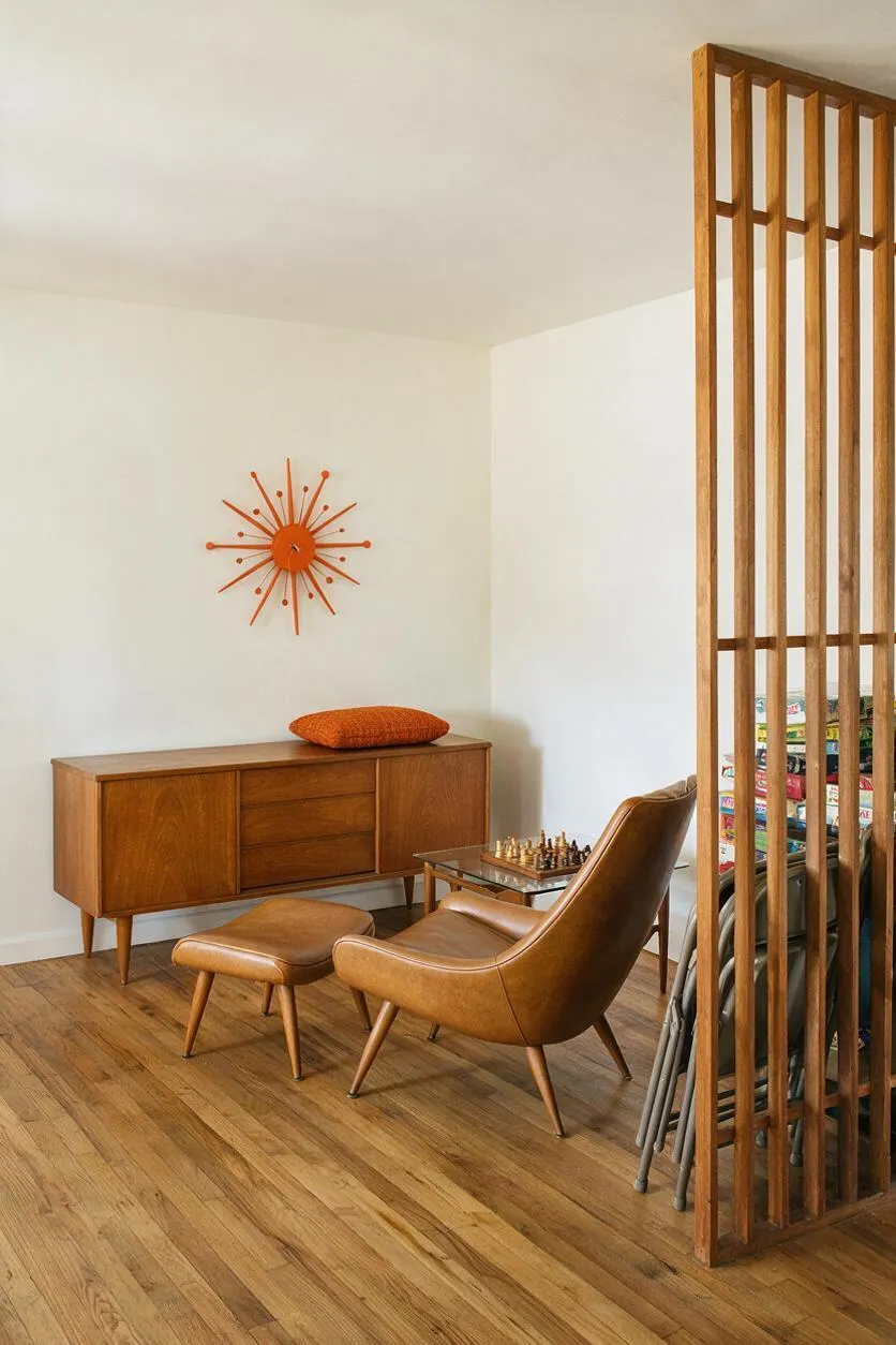 A mid-century modern game room with a walnut sideboard, tan leather lounge chair and ottoman, a small chess table, and an orange starburst wall clock. White walls and light wood floors keep the room bright, while a vertical wood slat partition adds architectural warmth and separates stacked games in the background.