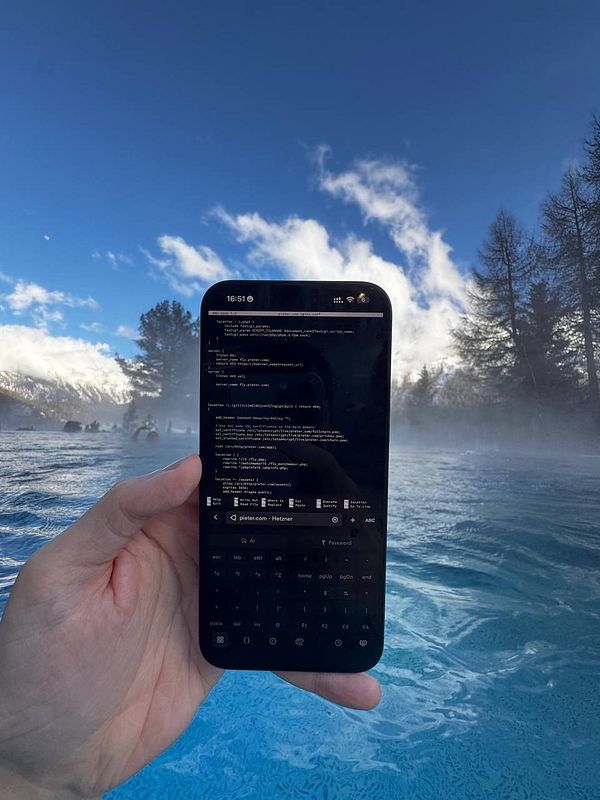 A hand holds a smartphone displaying code related to Nginx configuration against a scenic backdrop of a hot spring and mountains.