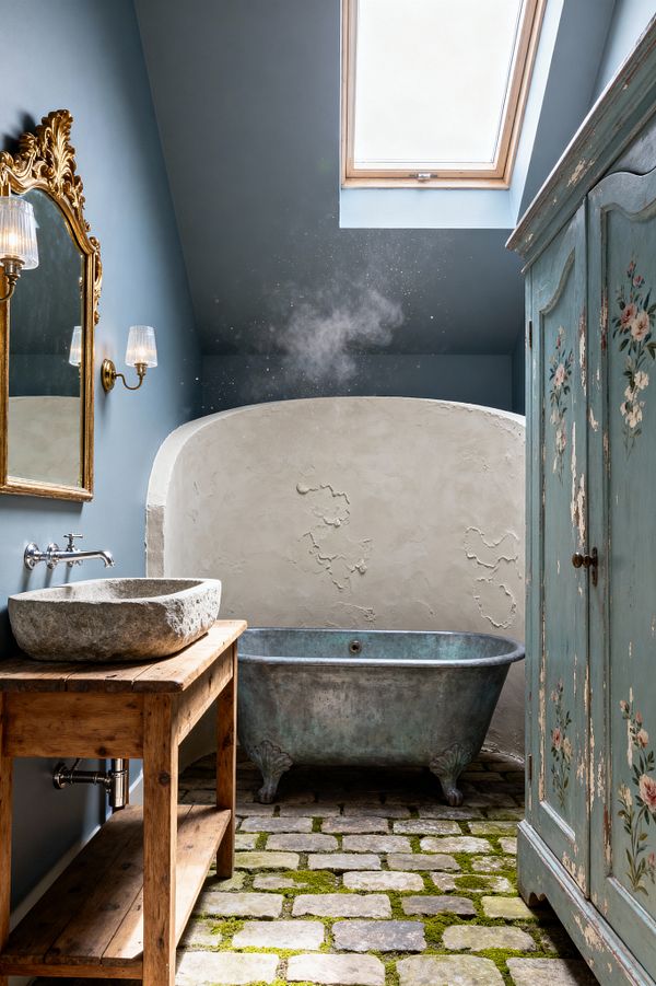 A French Country bathroom features a weathered zinc claw-foot tub before a curved, textured lime-plaster wall beneath a skylight. At left, a rustic pine console supports a rough-hewn stone vessel sink with a wall-mounted faucet, topped by an ornate gilded mirror and glass sconces. The floor is uneven limestone cobblestones with patches of moss. On the right stands a distressed blue-green armoire with hand-painted floral details, and the walls are painted dusty blue.